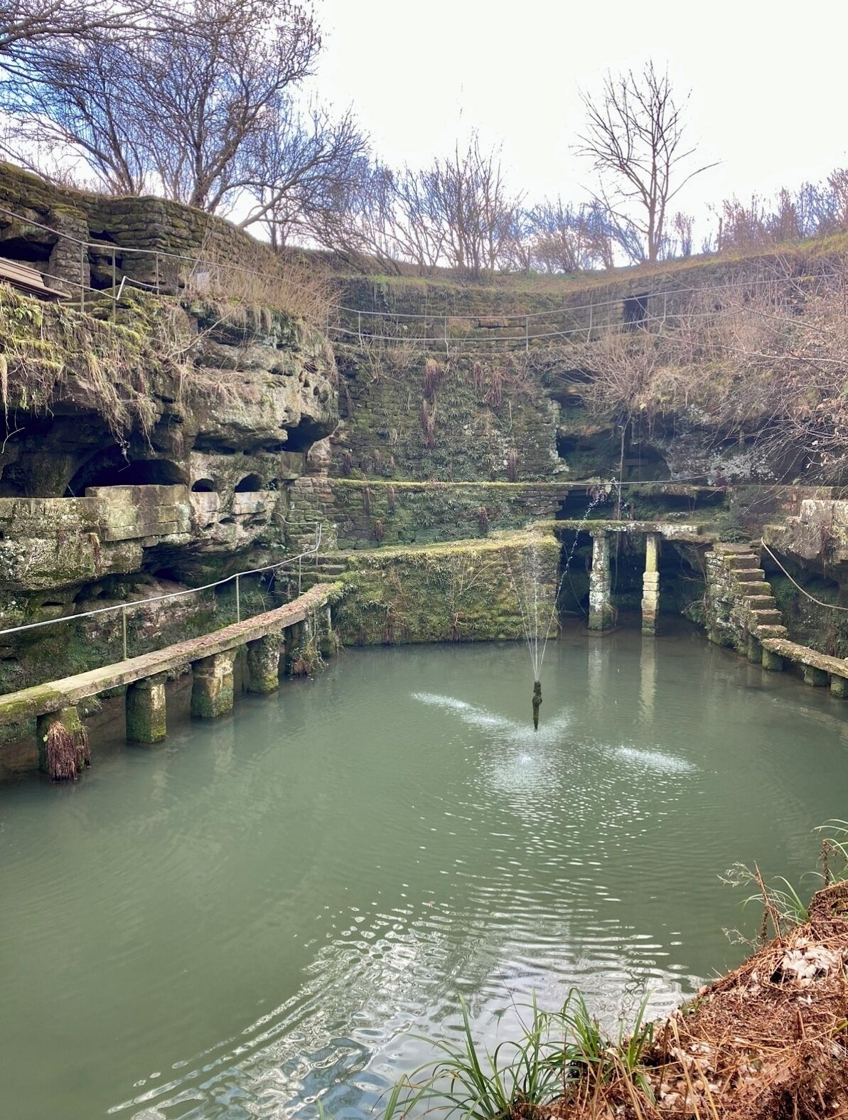 Un pont de pierre enjambant une étendue d’eau calme, avec des murs couverts de mousse et des arbres en arrière-plan.