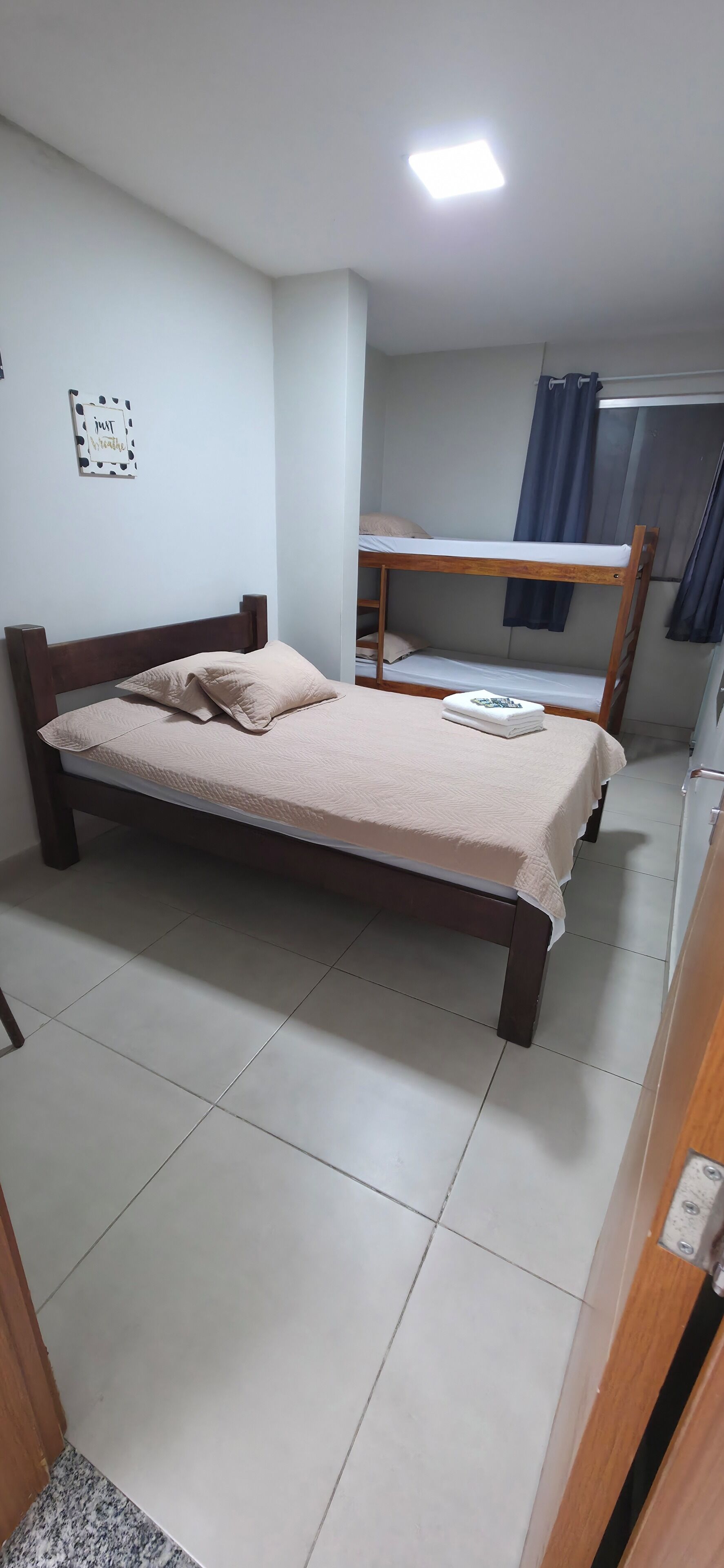 A single bed with a wooden frame and a beige bedspread, a bunk bed in the background, and a ceiling light.