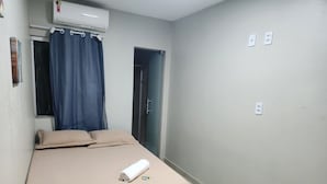 A small, single-bed room with a beige bedspread, a rolled towel, and a window with blue curtains. - Hangar House Hostel (Belém)
