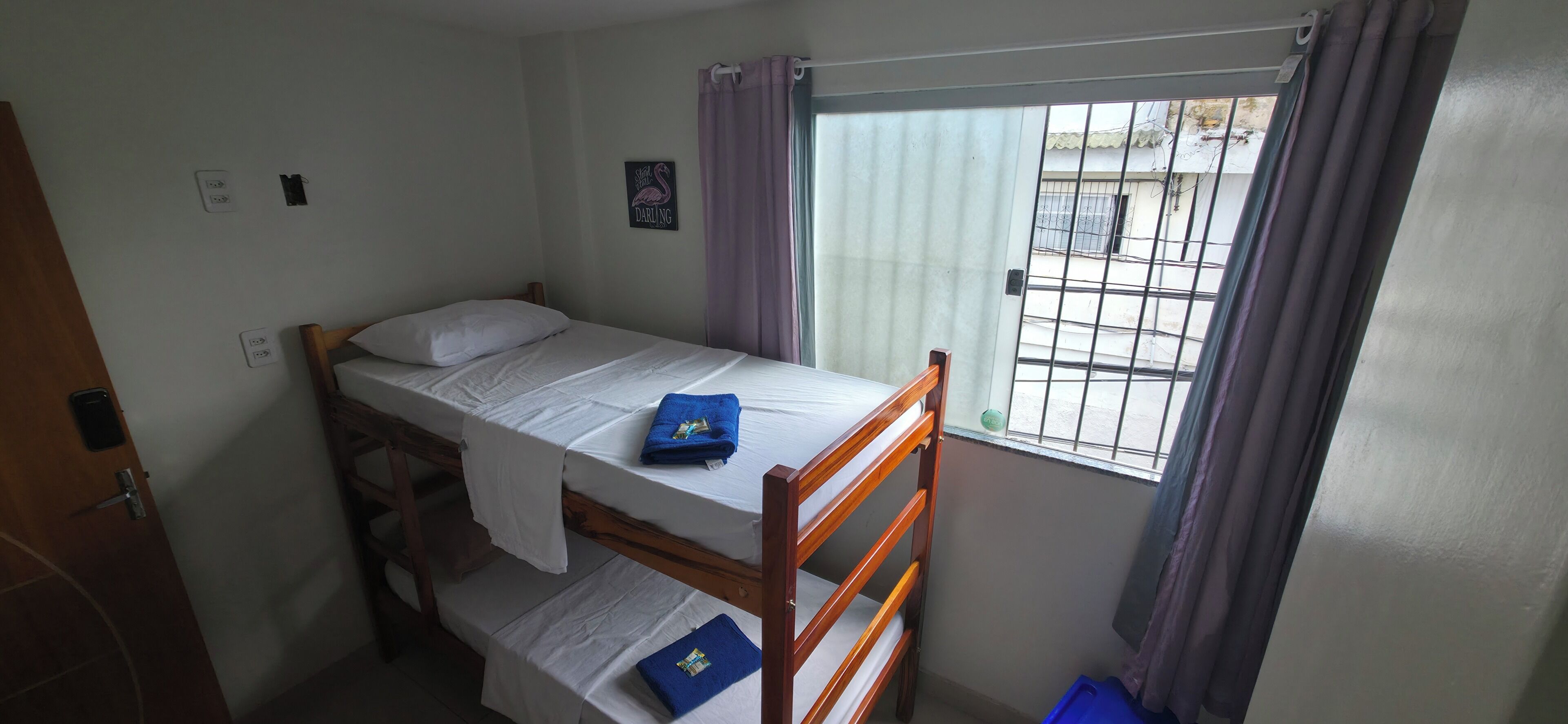 Una cama individual con estructura de madera, una ventana con barrotes metálicos y un interruptor de luz pequeño fijado a la pared.