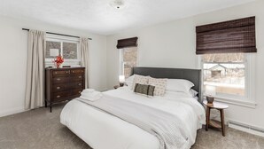 A bedroom with a large bed, a dresser, a nightstand, and two windows with blinds.
