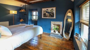 Un dormitorio con cama, suelo de madera, chimenea, espejo y ventana con persianas.