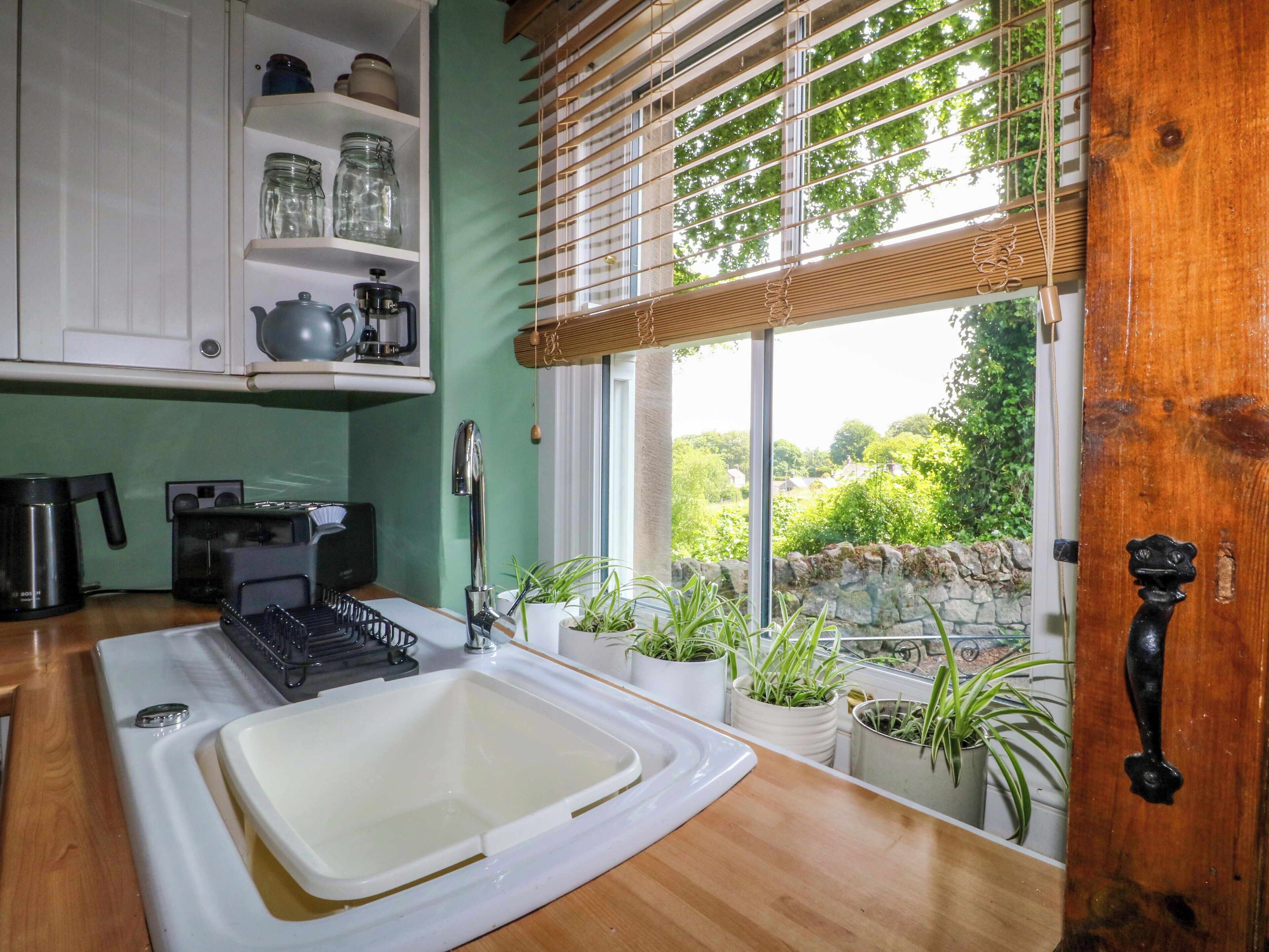 Une cuisine équipée d’un évier, d’une cuisinière et de divers appareils électroménagers. Des plantes en pot sont disposées sur le rebord de la fenêtre, et on peut admirer la verdure à l’extérieur.