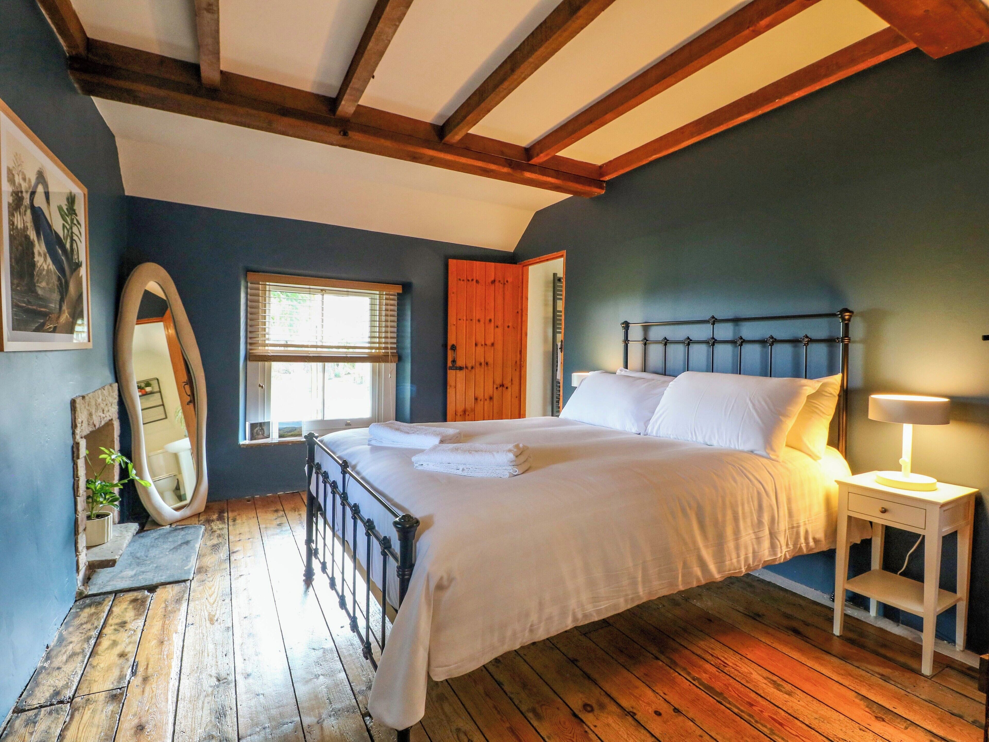 Un dormitorio con piso de madera, una cama con ropa blanca, una mesita de noche con lámpara, una puerta de madera y una ventana con persianas.