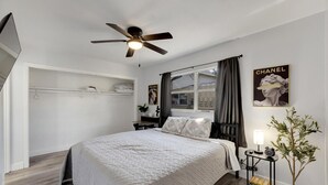 Une chambre à coucher comprenant un lit, un ventilateur de plafond, une fenêtre avec des rideaux, une table de chevet, une lampe et une plante en pot.