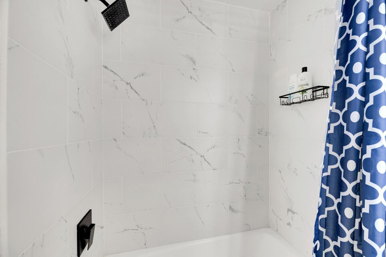 A bathroom with white marble tiles, a black showerhead, a white bathtub, and a blue patterned shower curtain.