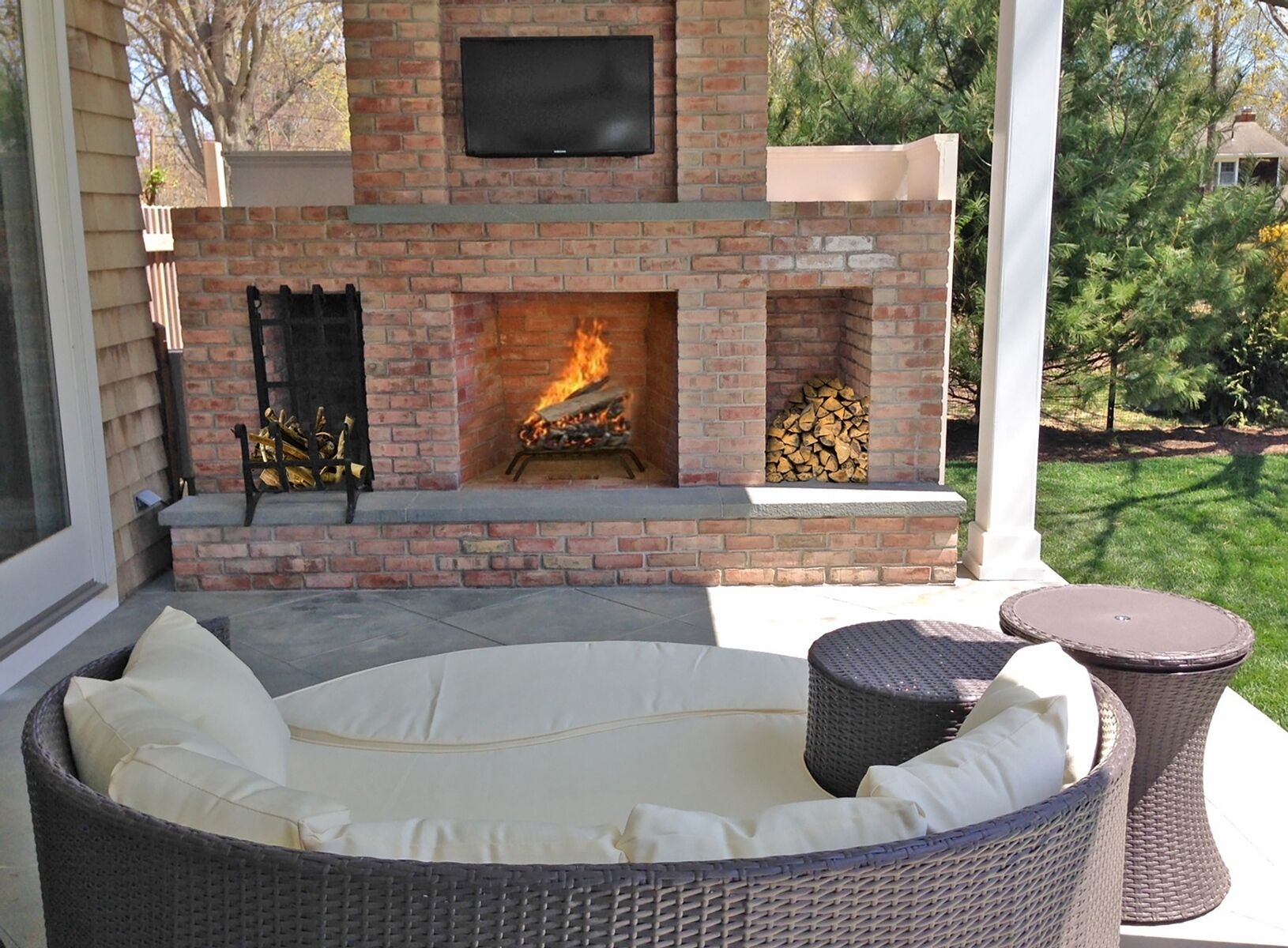 Eine Terrasse im Freien mit Kamin aus Backstein, einer gemütlichen Sitzecke und einem Fernseher, der über dem Kamin angebracht ist.