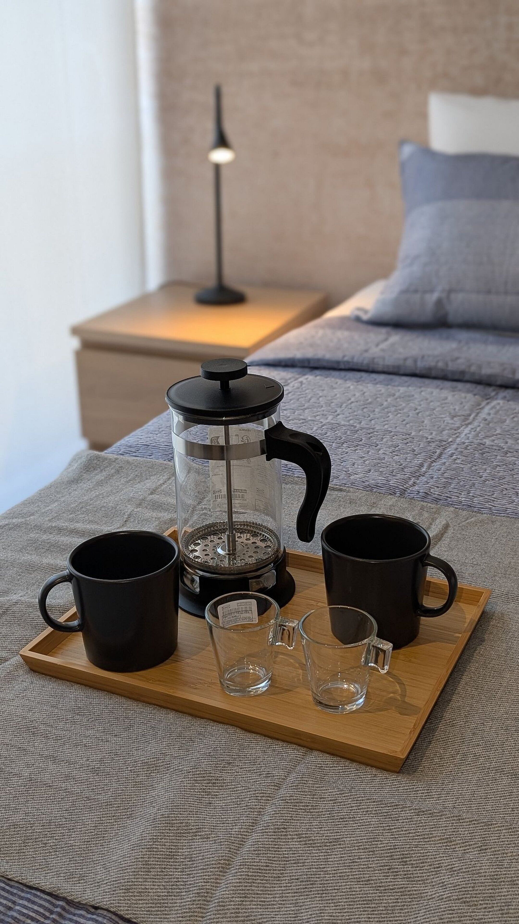 En kaffebord med en fransk stempelkande, to sorte krus og to glas på en seng med et gråt tæppe, og en lampe i baggrunden.
