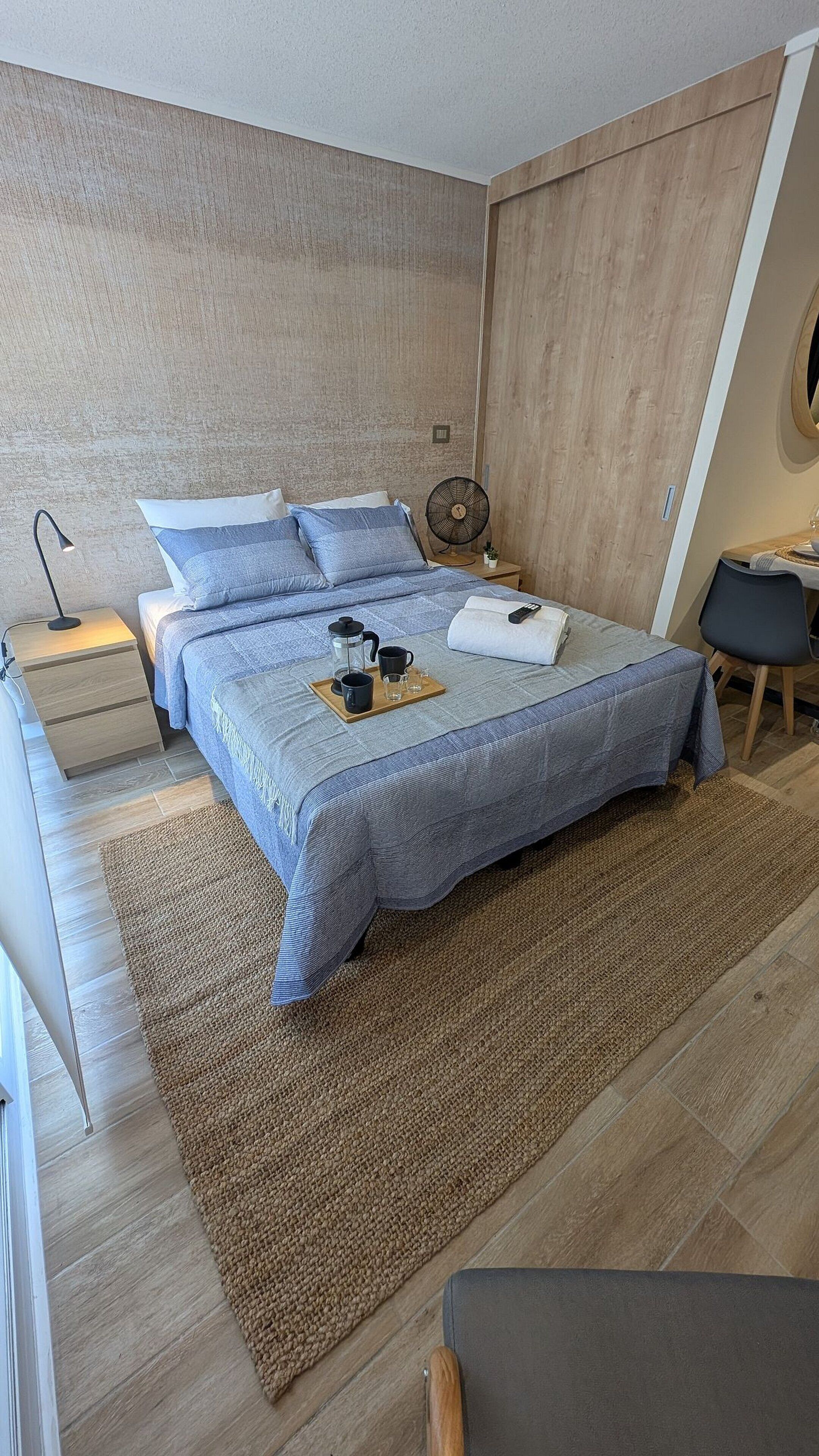 A modern bedroom with a bed, bedside table, lamp, and a small table with a fan and a remote control.