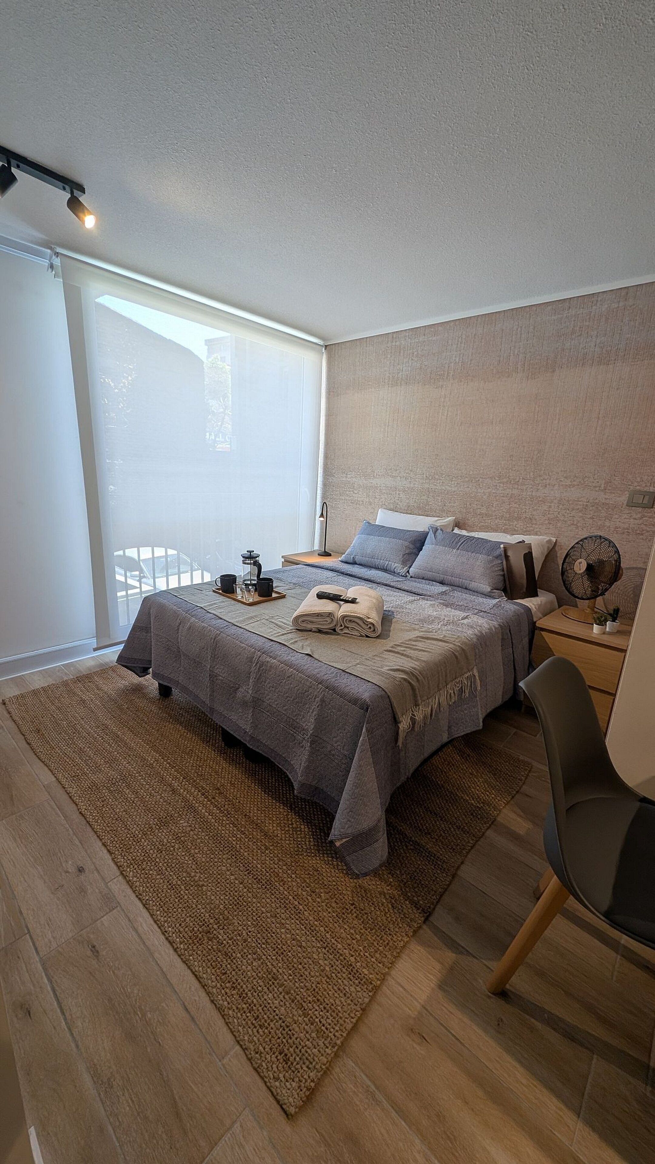 Un dormitorio moderno con una cama grande, una mesita de noche con lámpara, una silla y vistas al exterior a través de un ventanal.