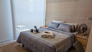 Un dormitorio moderno con una cama grande, una mesita de noche con lámpara, una silla y vistas al exterior a través de un ventanal.