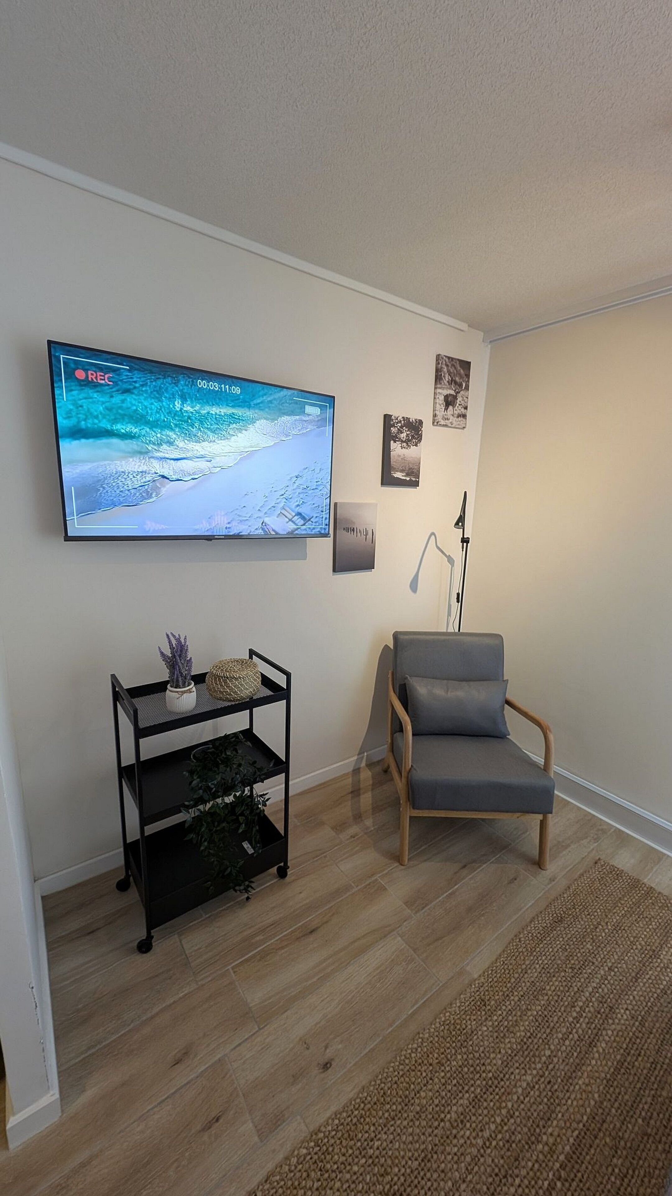 Un salon avec un téléviseur à écran plat fixé au mur, un fauteuil gris, une étagère noire ornée d’objets décoratifs et un lampadaire.