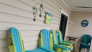 A row of colorful Adirondack chairs on a wooden deck with a white exterior wall, a small table, and a door.
