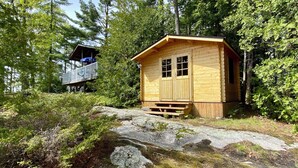 Pieni puinen mökki, jossa on pieni kuisti ja parveke, ja sen ympärillä on tiheää metsää.