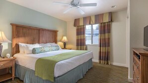 A hotel room with a bed, two bedside tables with lamps, a ceiling fan, a window with curtains, and a TV on a wooden stand.
