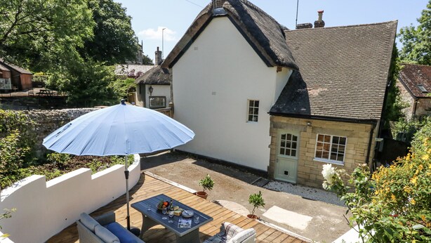 A patio with a wicker sofa, a table with a bottle and glasses, and a blue umbrella. - Happiness Cottage (Dorchester)