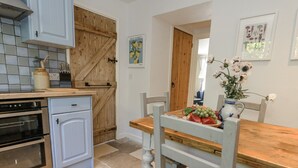 A kitchen with a wooden door, a dining table with a vase of flowers, and a fruit bowl. - Happiness Cottage (Dorchester)