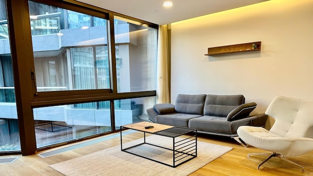 Uma sala de estar moderna com um sofá cinza, uma poltrona branca, uma mesa de centro de madeira e uma grande janela com vista para o exterior do edifício.