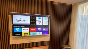 Uma sala de estar moderna com uma TV fixada na parede exibindo diversos aplicativos de streaming, uma parede com ripas de madeira, um pequeno banquinho de madeira e uma obra de arte emoldurada abaixo da TV.