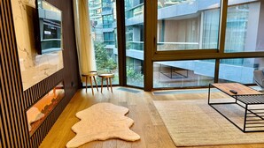 Uma sala de estar moderna com uma janela ampla, piso de madeira, um banquinho de couro e uma mesa pequena.