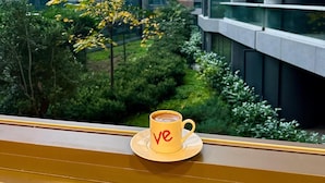Uma caneca amarela com texto vermelho numa janela com vista para um edifício moderno de vidro e concreto.