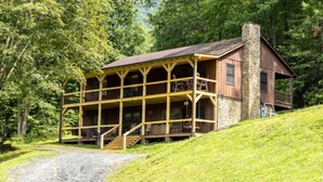 Eine Blockhütte mit Veranda und Kamin aus Stein, umgeben von üppigem Grün.