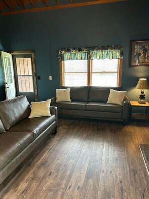 A living room with a dark brown leather sofa, a matching armchair, and a wooden coffee table. There is a window with blinds and a curtain, a door with a window, and a framed picture on the wall. - The River View Cabin (Spruce Pine)