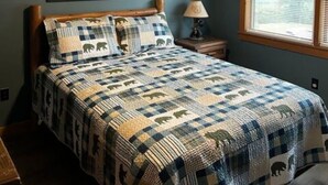 A bedroom with a wooden ceiling, a bed with a quilt featuring animal prints, a framed picture on the wall, and a window with a checkered curtain. - The River View Cabin (Spruce Pine)
