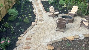 Ein Garten mit Kiesweg, Holzzaun und Sitzbereich im Freien mit Stühlen und Feuerstelle.