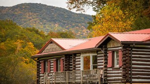 A log cabin with a porch and a stone foundation, surrounded by autumn foliage and a mountain in the background. - The Valley Lodge Sleeps 12 (Spruce Pine)