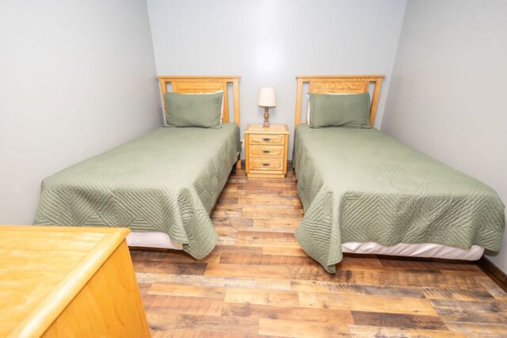 Een kleine, schone kamer met twee eenpersoonsbedden, een houten nachtkastje met lamp en een houten dressoir.