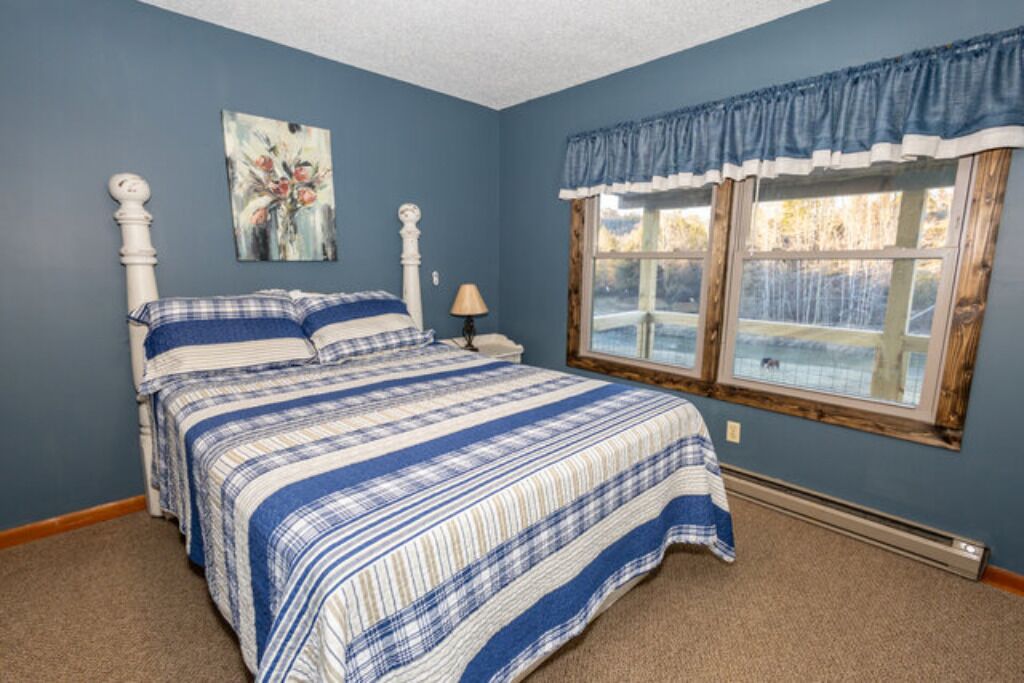 A bedroom with a bed, a nightstand, a lamp, a window with curtains, and a painting on the wall.