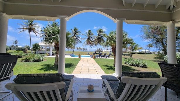 A covered outdoor seating area with white chairs and a table, overlooking a pool and palm trees.