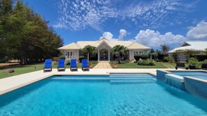 A swimming pool with blue water, surrounded by a white deck. There are three blue lounge chairs and a small patio area with a table and chairs.