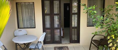 A small outdoor patio area with a table and chairs, a door leading inside, and some potted plants.