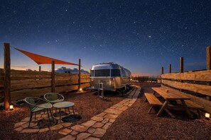 A starry night sky with a vintage trailer and picnic tables in a gravel area.