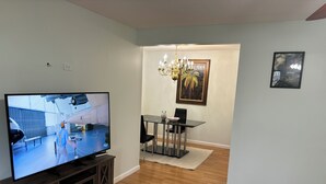 Un salon moderne doté d’un téléviseur à écran plat, de planchers en bois, d’un coin repas avec une table et des chaises en verre, d’un lustre et d’un tableau encadré au mur.