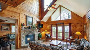 A cozy cabin interior with a stone fireplace, wooden furniture, and large windows.