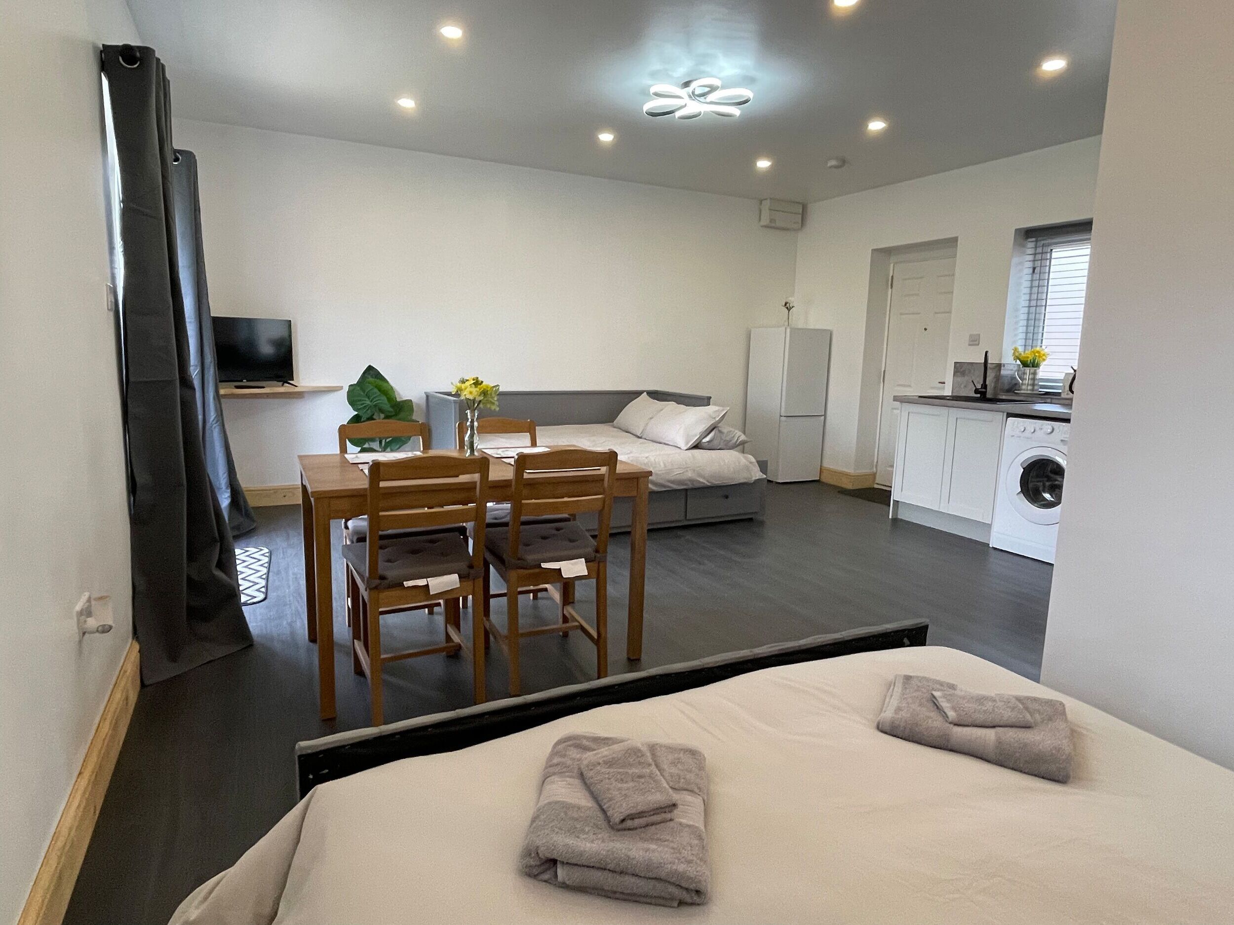 A modern hotel room with a bed, a small dining table with chairs, a sofa, a TV, and a kitchen area with a washing machine.