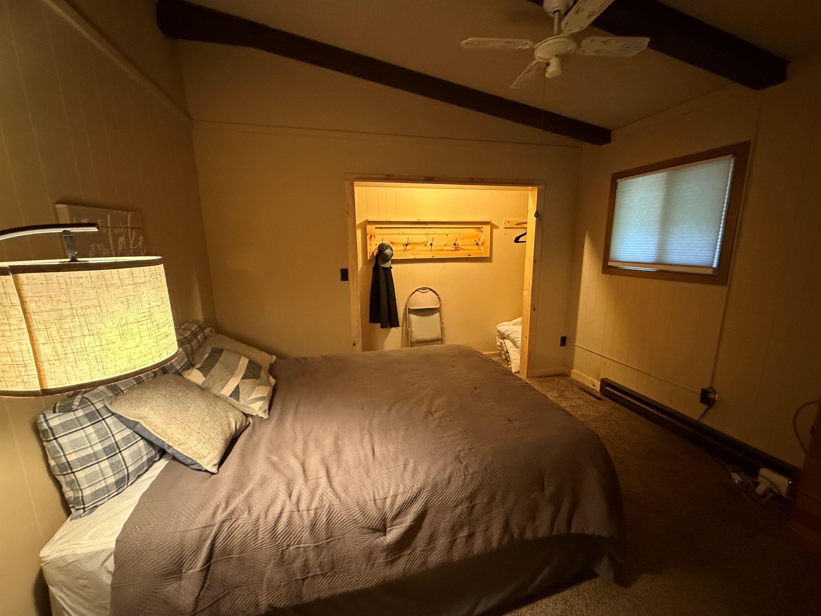 Un dormitorio con cama, mesita de noche, lámpara, ventilador de techo, ventana con persianas y puerta que da a otra habitación.