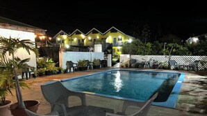 Piscina com uma pequena mesa e cadeiras em primeiro plano, cercada por plantas em vasos e um muro branco.