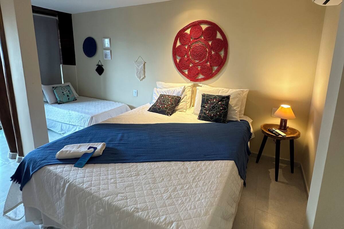 Ein Hotelzimmer mit einem großen Bett, einem kleineren Bett, einem Wandbild, einer Lampe und einer Fernbedienung auf einem Nachttisch.
