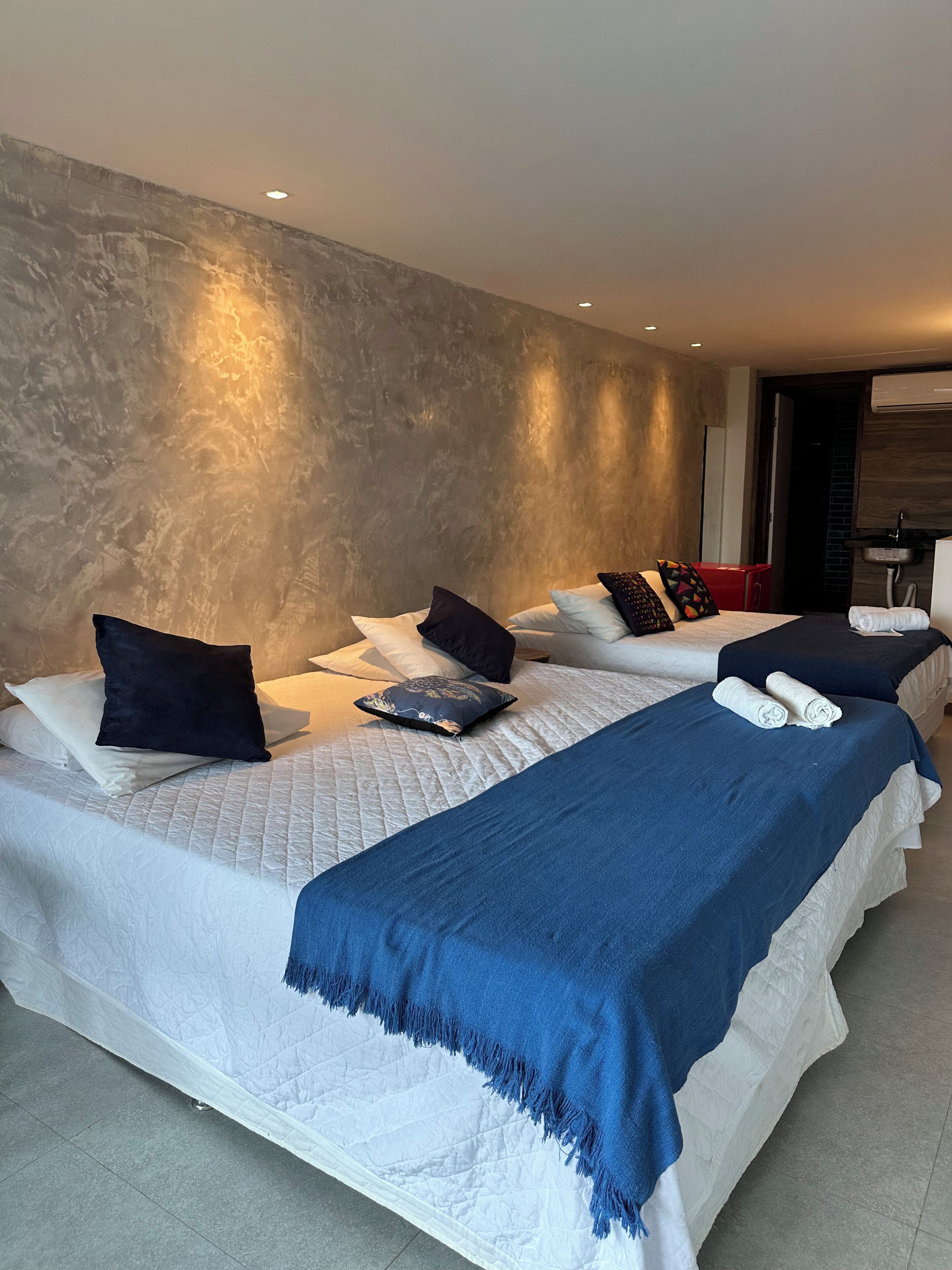 A hotel room with three beds, each with a white quilted bedspread and blue throw blankets. The room has a textured gray wall and a modern air conditioning unit mounted on the wall.