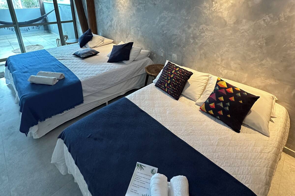 Ein Hotelzimmer mit zwei Betten, jeweils mit blauer und weißer Bettdecke, und einem farbenfrohen geometrischen Kissen. In der Ecke steht ein kleiner Tisch mit einem Stuhl, und ein großes Fenster bietet einen Blick nach draußen.