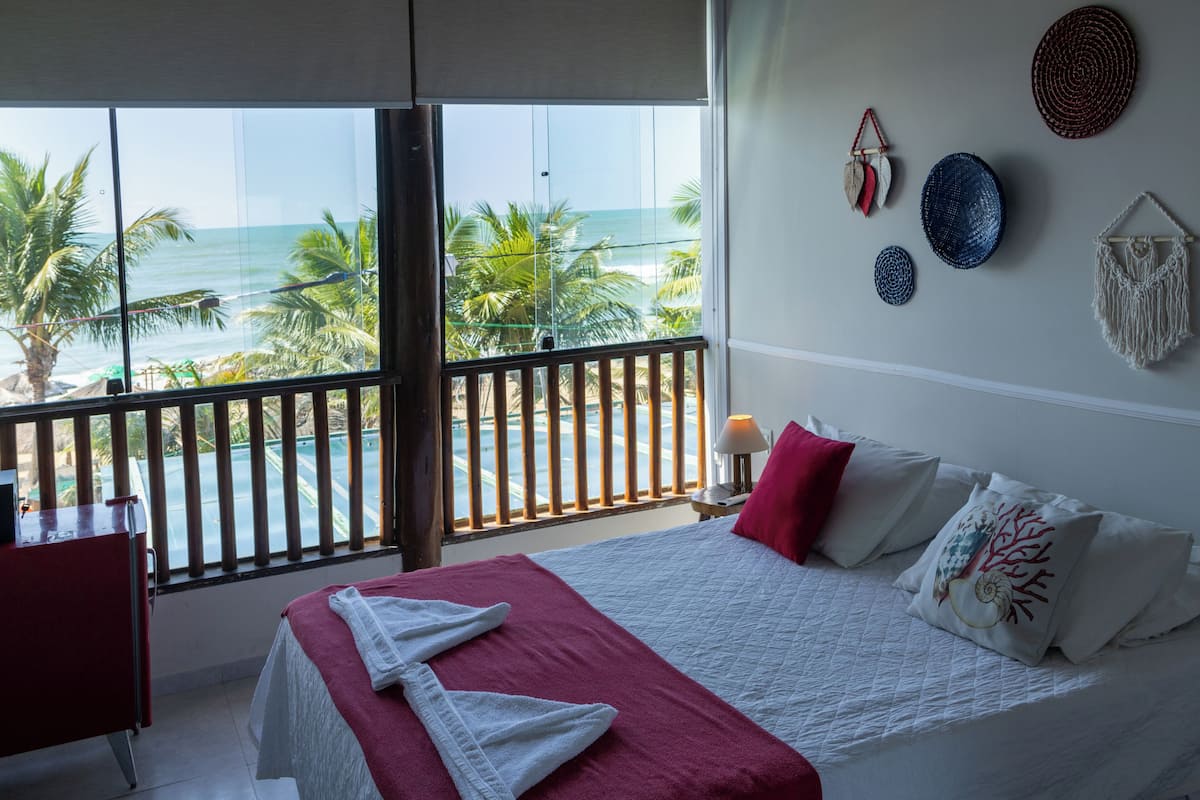 Ein Schlafzimmer mit großem Fenster mit Blick auf den Ozean, einem Bett mit weißer und roter Bettwäsche sowie dekorativen Wandgestaltungen.