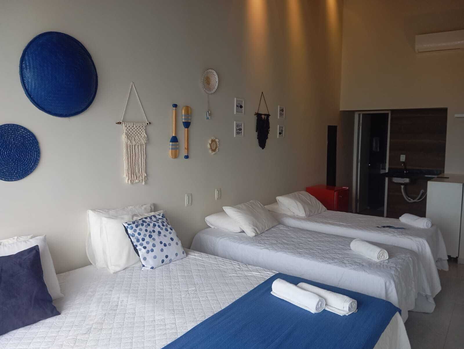 Quarto de hotel com duas camas, um tapete azul e decorações na parede, incluindo um pendente de macramé e objetos de madeira.