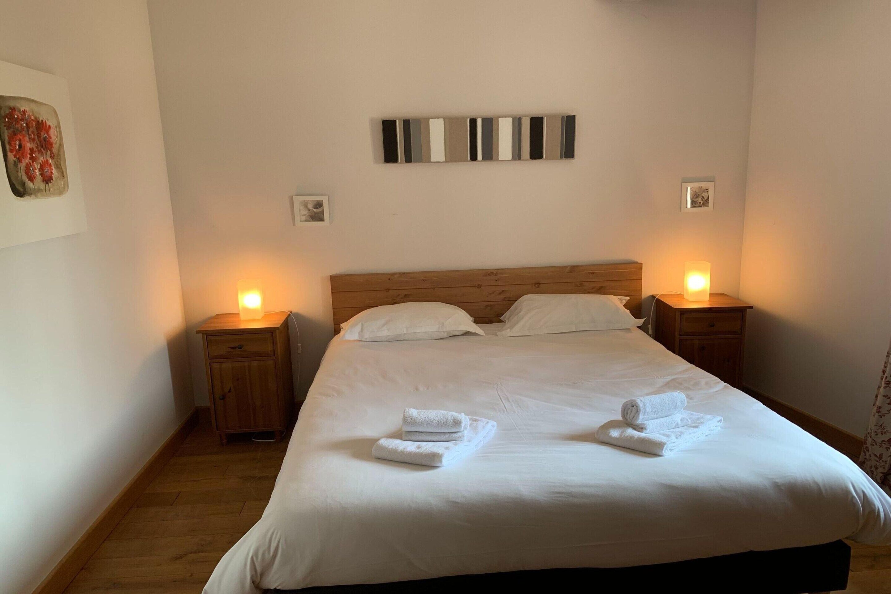 A neatly made bed with white linens and two folded towels on the bed. There are two bedside tables with lamps, and the room has a wooden floor.