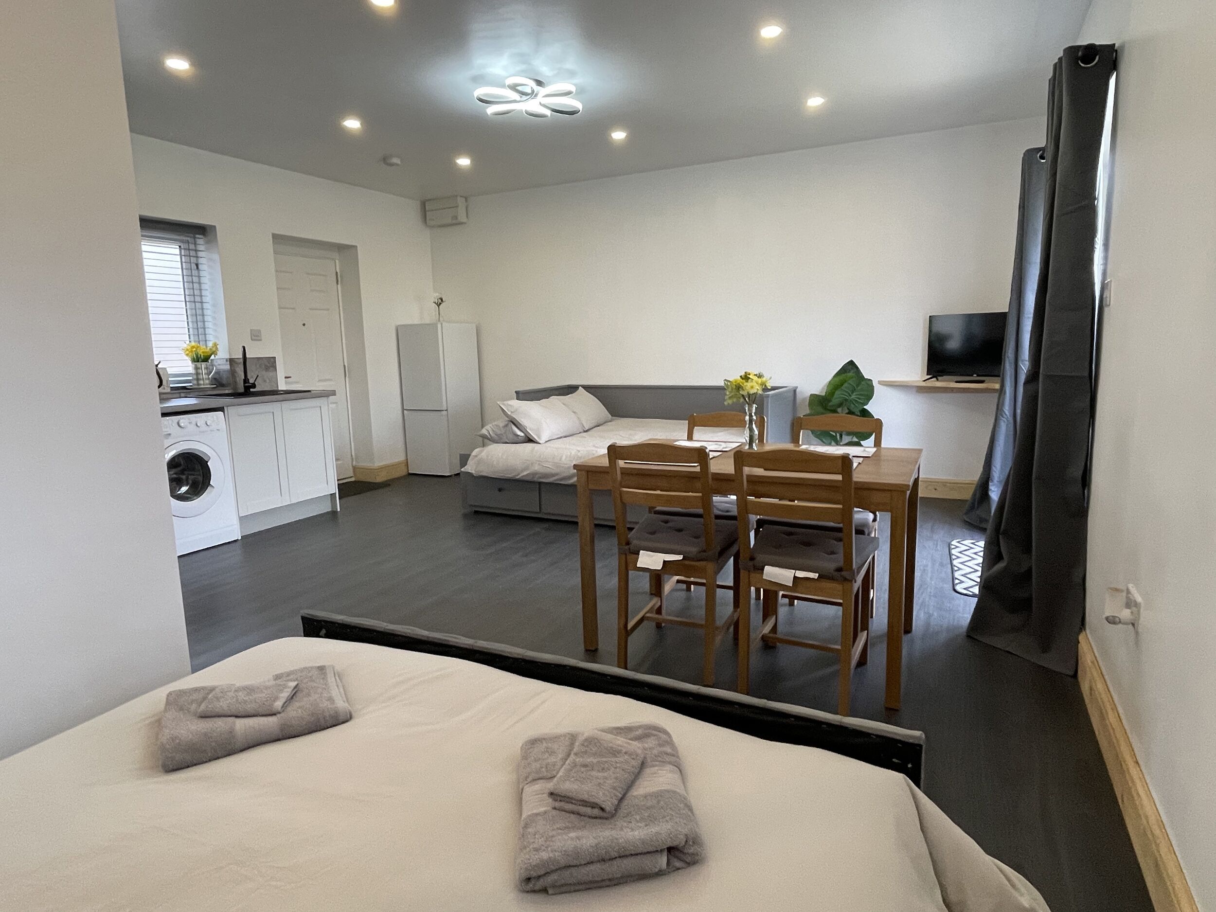 Habitación de hotel moderna con cama, mesa de comedor con sillas, televisión y cocina equipada con lavadora.