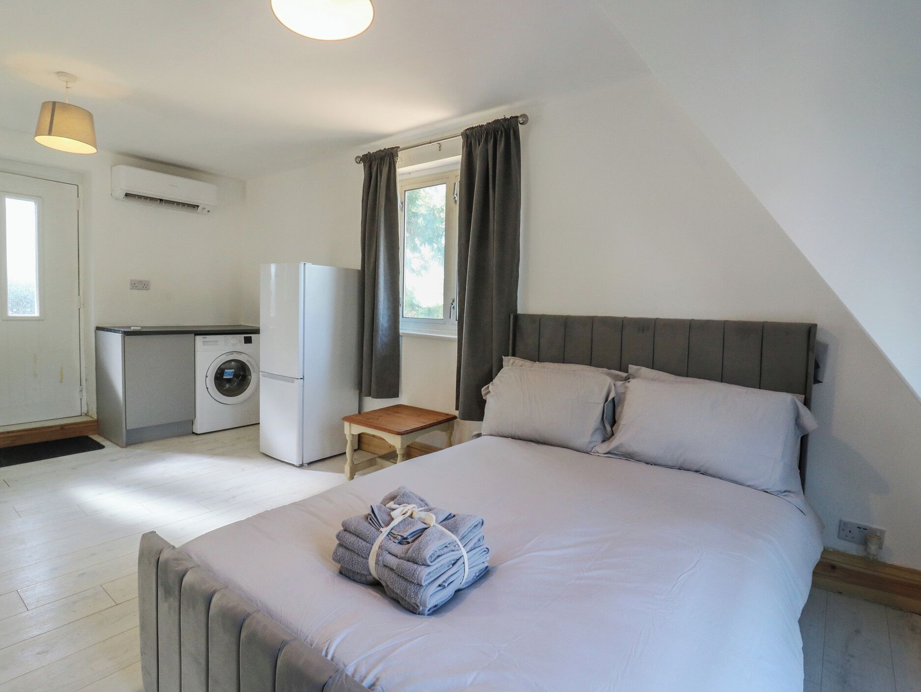 Een compacte slaapkamer met een bed, een klein tafeltje en een kitchenette, inclusief een wasmachine en een koelkast.