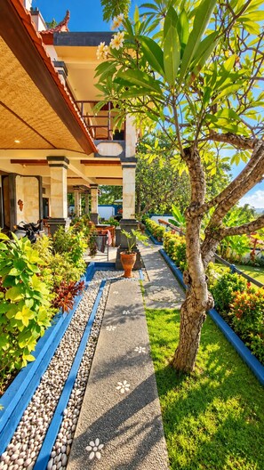 A pathway lined with potted plants and a tree, leading to a building with a tiled roof and outdoor seating area. - Stunning 3-Bedroom Hilltop Villa with Ocean Views - Dejaviews Villa (Bali)